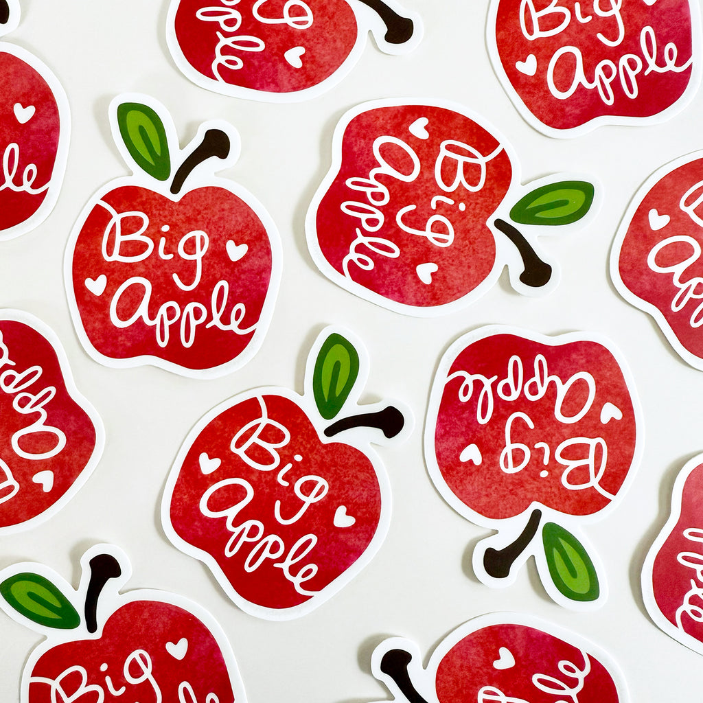 A group of 12 apple-shaped New York City-themed Big Apple vinyl stickers are shown scattered across a white background. Featuring hand lettered Big Apple text and 2 white hearts, each NYC inspired waterproof souvenir sticker is made in the USA.