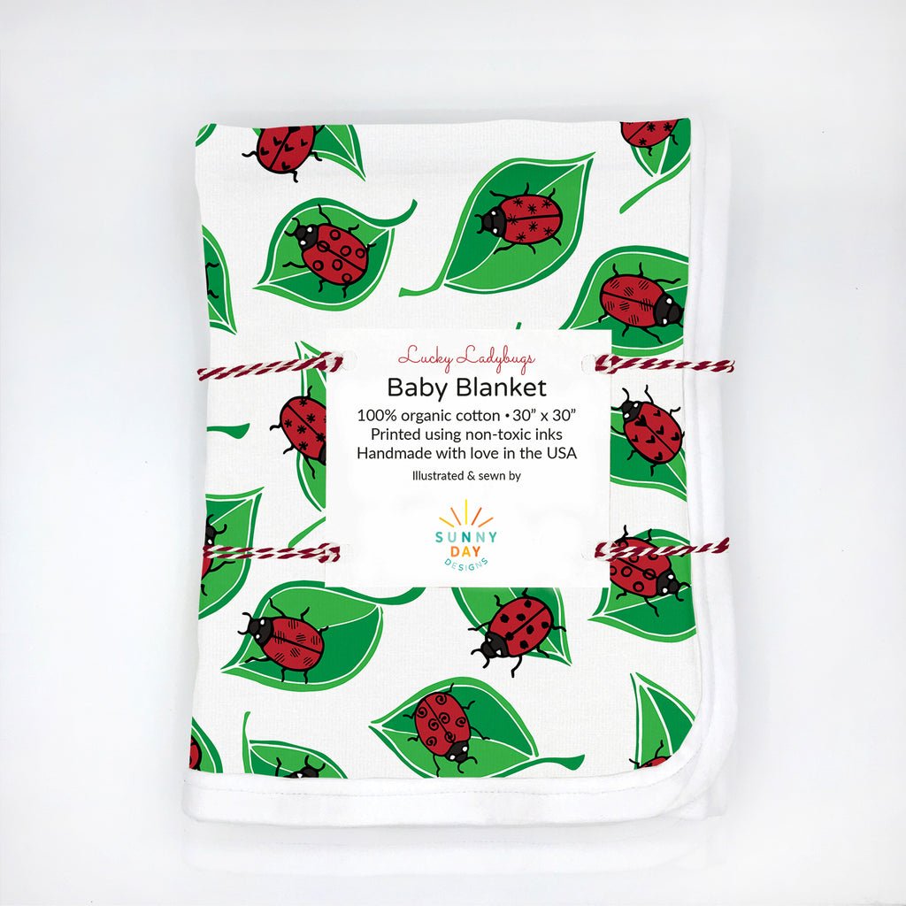An organic cotton baby blanket is shown packaged and features a red, black, green, and white ladybug and leaf print design. Made in the USA by Sunny Day Designs.