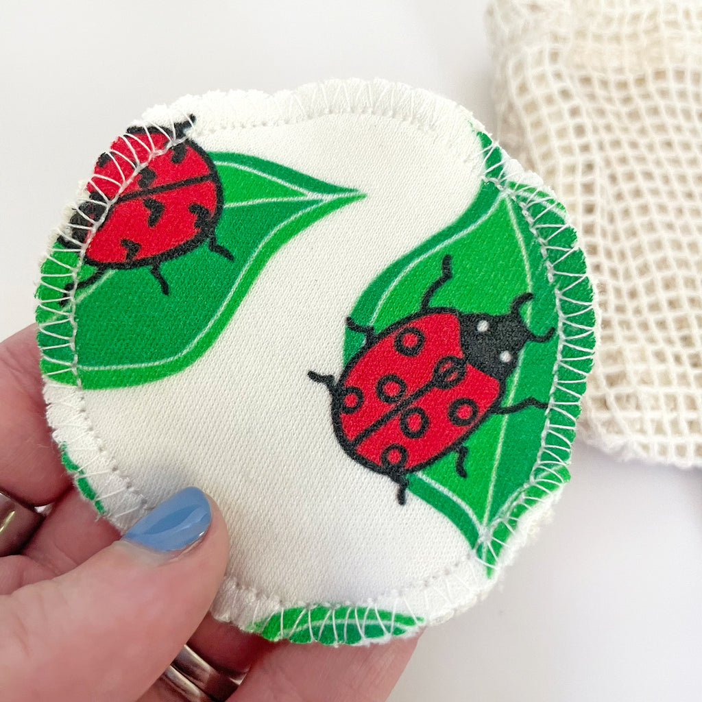 Close-up of a reusable facial round featuring a ladybug pattern, held in hand with an eco-friendly mesh wash bag in the background.