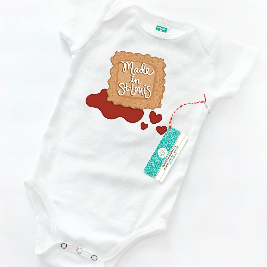 A white organic cotton baby onesie by Sunny Day Designs is shown lying on top of a white background and printed with a Saint Louis food-themed design showing a tan/brown toasted ravioli with the words" Made in St. Louis" written on it in hand lettered script, and sitting in a puddle of reddish-orange marinara sauce with 3 heart shaped drips.