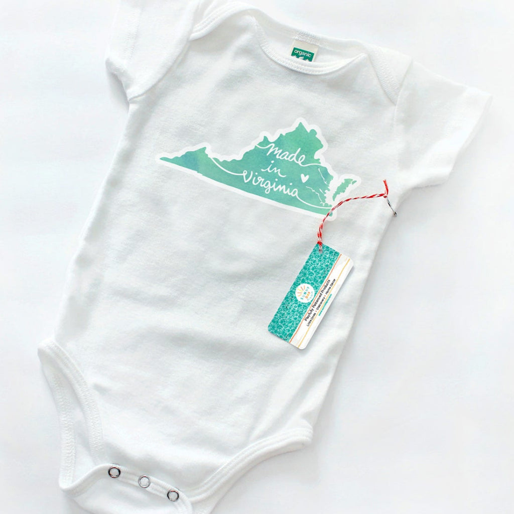 White baby onesie featuring a mint green silhouette of the state of Virginia with the phrase “Made in Virginia” in white script font. Displayed flat on a clean white background with a red and white string hang tag labeled Sunny Day Designs, showcasing minimalist eco-conscious branding.