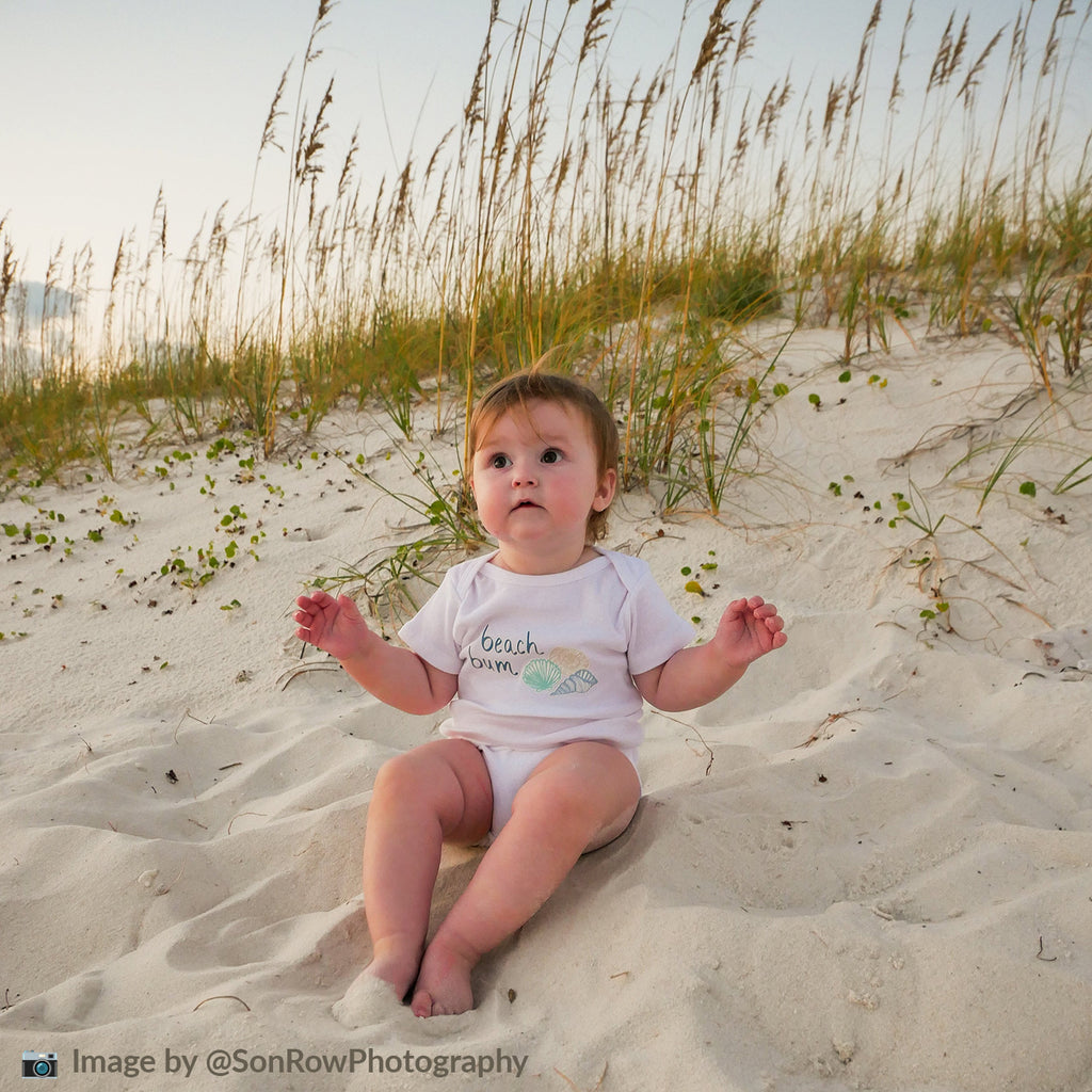 Baby sitting on the sand at the beach wearing a seashell themed white onesie that says "beach bum". Photo by SonRow Photography and taken on the Florida panhandle.