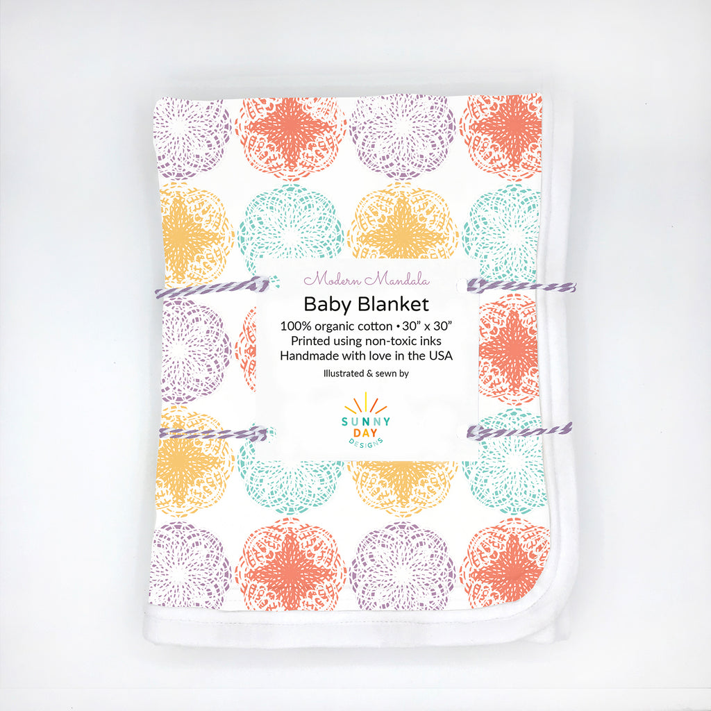 Folded organic cotton baby blanket with a modern mandala print in vibrant pastel tones orange, purple, mint green, and golden yellow on a white background. Packaged with a white label that reads “Modern Mandala Baby Blanket – 100% organic cotton, 30” x 30”, printed using non-toxic inks, handmade with love in the USA.” Flat lay style with eco-conscious branding by Sunny Day Designs.