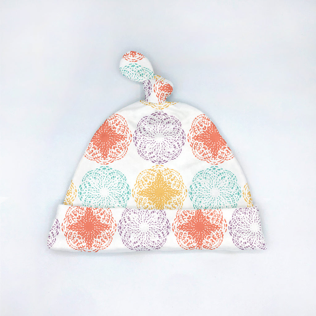 Colorful baby hat with a top knot featuring a repeating mandala-style circular pattern in pastel orange, mint, yellow, lavender purple & white. Displayed flat on a white background, from the Modern Mandala eco-friendly baby line by Sunny Day Designs.