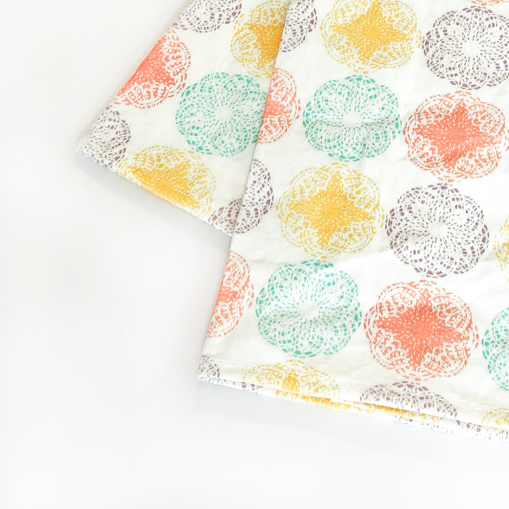 Angled close-up view of the folded Modern Mandala Tea Towel. The vibrant circular designs in warm shades of yellow, orange, teal, and purple stand out against the white background, emphasizing the towel’s contemporary, artistic look.