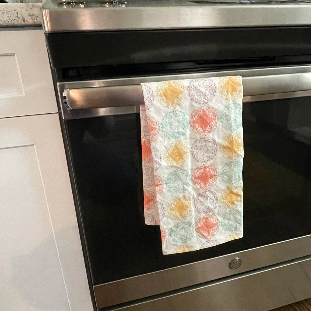 Modern Mandala Tea Towel hanging from a stainless steel oven door handle in a white kitchen setting. The towel’s bright mandala pattern in yellow, teal, orange, and purple adds a vibrant touch to the kitchen décor while being practical for everyday use.