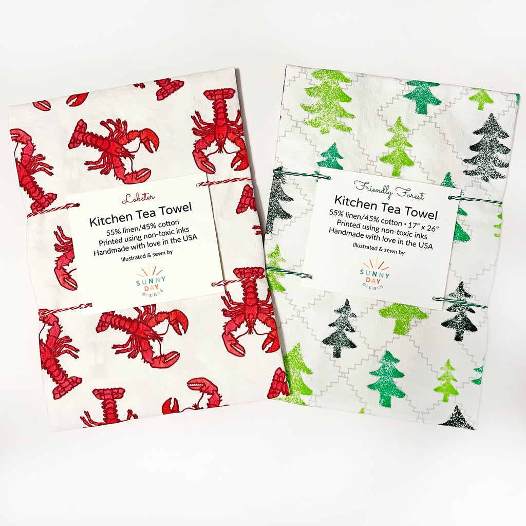 A cute set of 2 colorful New England-themed kitchen tea towels by Sunny Day Designs (1 in a red lobster print and 1 in a green tree/forest print) are shown packaged with colorful red and green baker's twine on a white background.