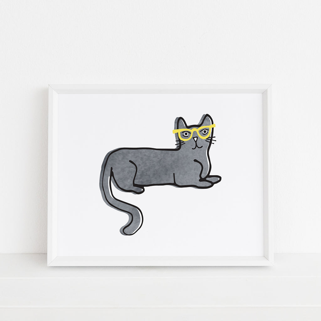 A quirky, framed illustration titled "One Cool Cat" by Sunny Day Designs - a gray watercolor cat who is smiling and wearing yellow glasses while laying down. Art print is framed in a white frame.