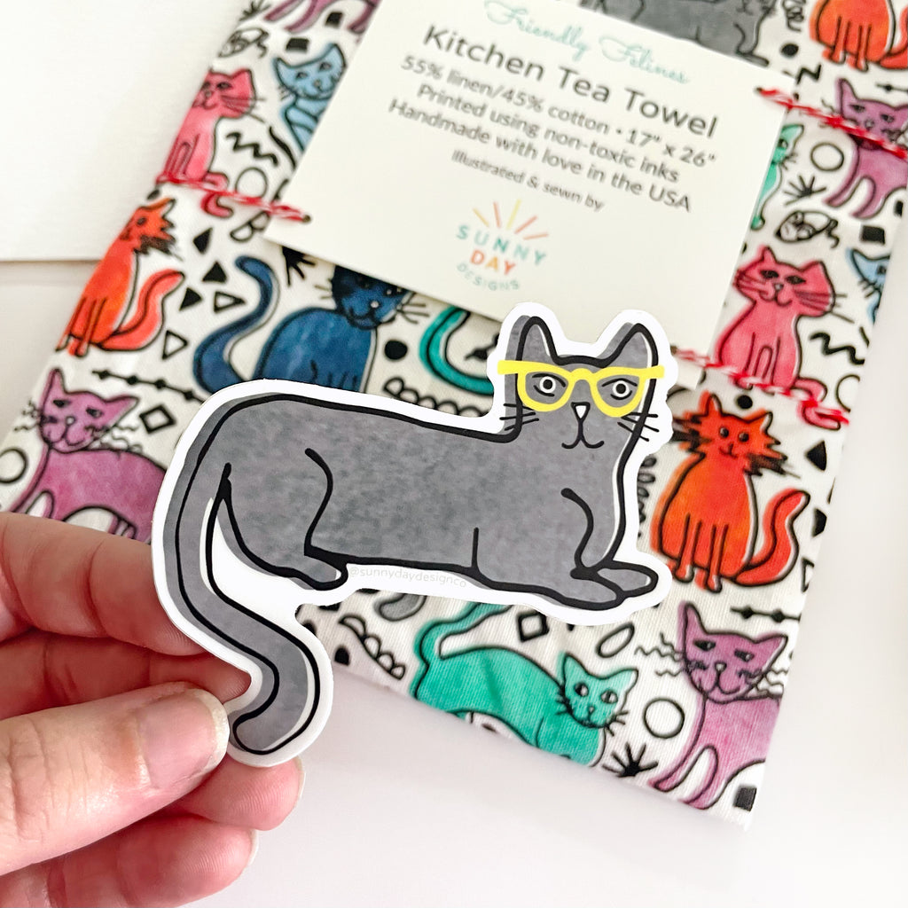A hand holds a cute watercolor vinyl sticker of a gray cat laying down and wearing bright yellow glasses while a colorful, cat-themed printed kitchen tea towel is shown packaged in the background. This cat printed dish towel is joyfully made in the USA from a durable and soft linen/cotton fabric that is custom printed for Sunny Day Designs.