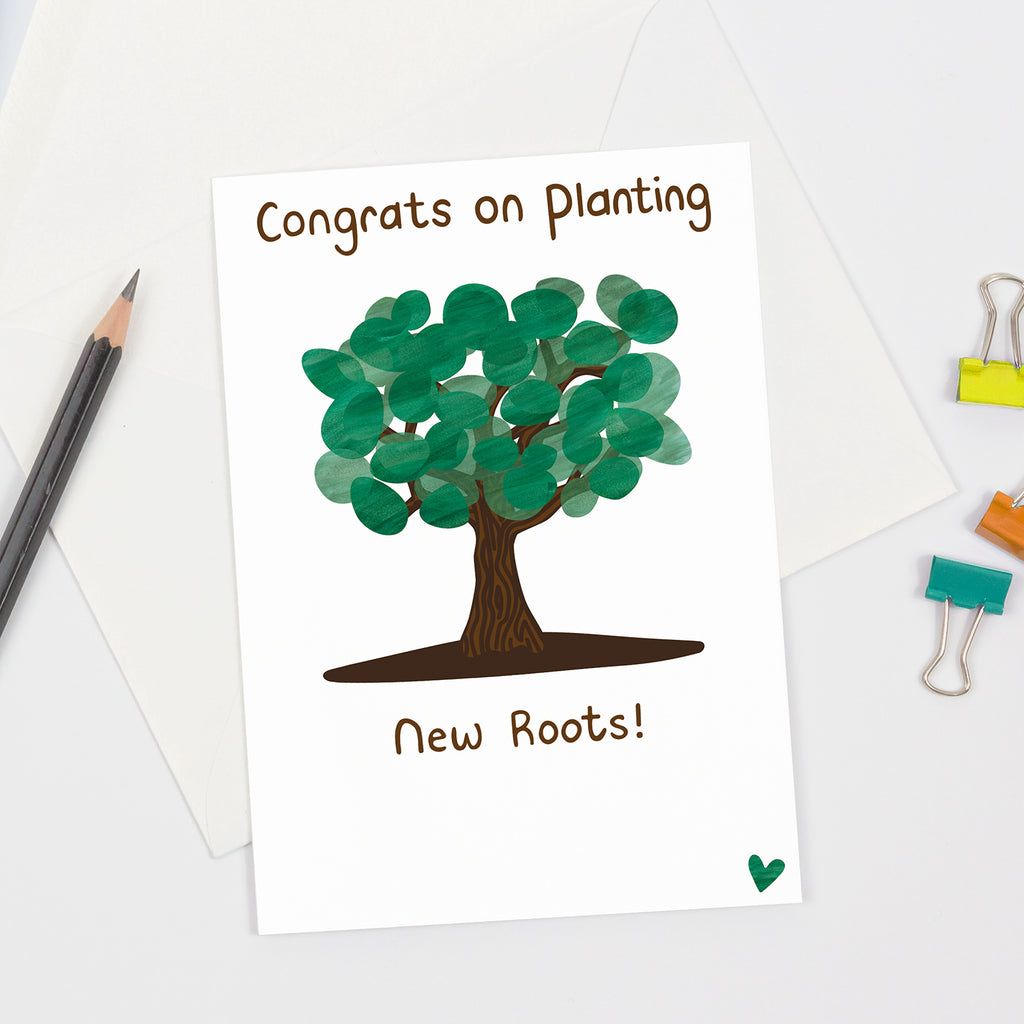 Greeting card featuring an illustrated tree with layered green leaves and brown trunk, and the message “Congrats on Planting New Roots!” in brown handwritten-style font. Flat lay on a white background with envelope, pencil, and colorful office accessories.