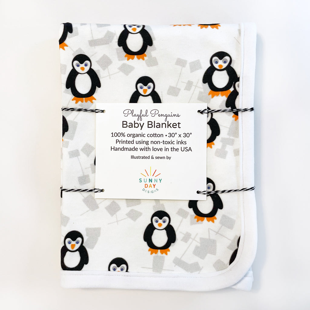 An adorable, handmade in the USA playful penguin-printed baby blanket by Sunny Day Designs is shown packaged with a card and black/white baker's twine and folded on top of a white background.