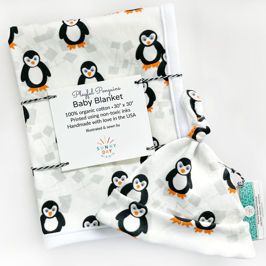 An organic cotton penguin themed newborn baby gift set by Sunny Day Designs is displayed and features a playful penguin printed baby blanket and penguin printed baby hat. Both items are shown packaged and folded on top of a white background.