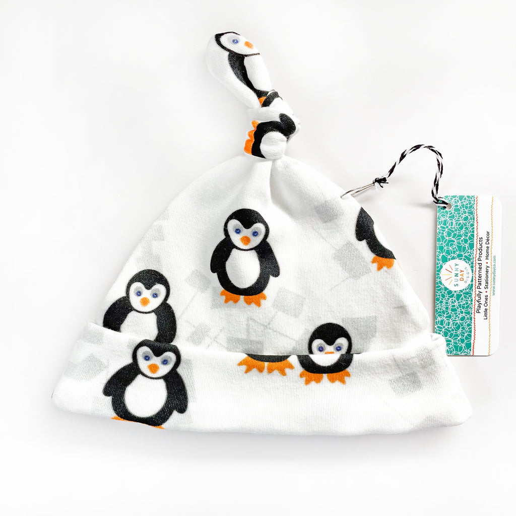 A black, white & orange penguin themed printed baby hat made from organic cotton by Sunny Day Designs is shown laying flat and packaged with a hang tag