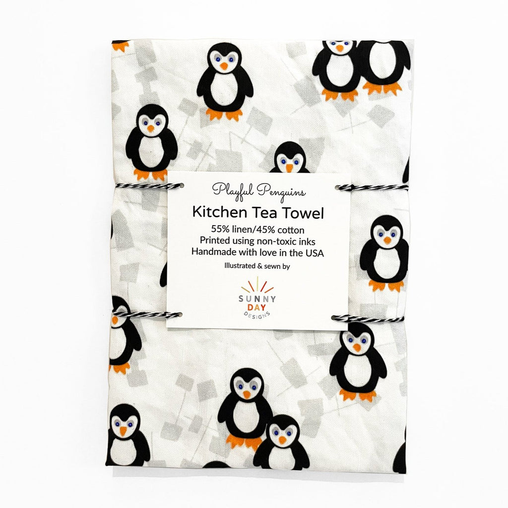 Playful Penguins kitchen tea towel, made of 55% linen and 45% cotton, featuring a whimsical penguin pattern on a white background. The towel is packaged with a label that reads 'Playful Penguins Kitchen Tea Towel' and includes product details like non-toxic inks and handmade in the USA by Sunny Day Designs.