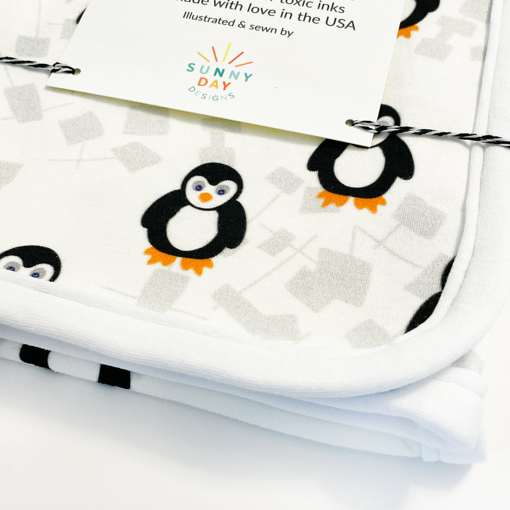 A close up image of a packaged organic cotton penguin printed baby blanket by Sunny Day Designs that shows the white organic cotton fabric binding and curved corners of this 30" square baby receiving blanket.