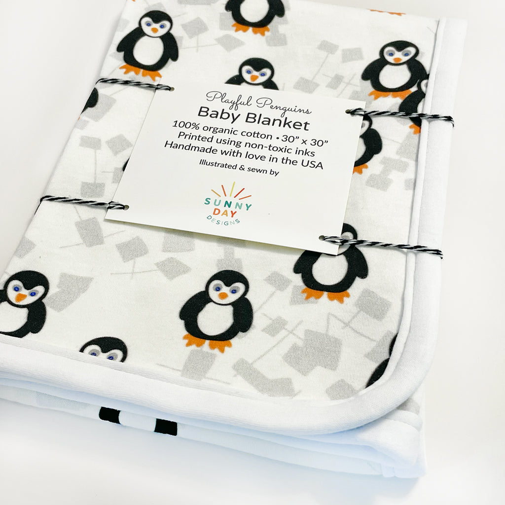 An adorable, handmade arctic animal-themed baby blanket by Sunny Day Designs is shown packaged with black baker's twine and features a cute black, white and orange playful penguin print design. This organic cotton baby blanket is made in the USA from super soft fabric printed with baby safe inks.