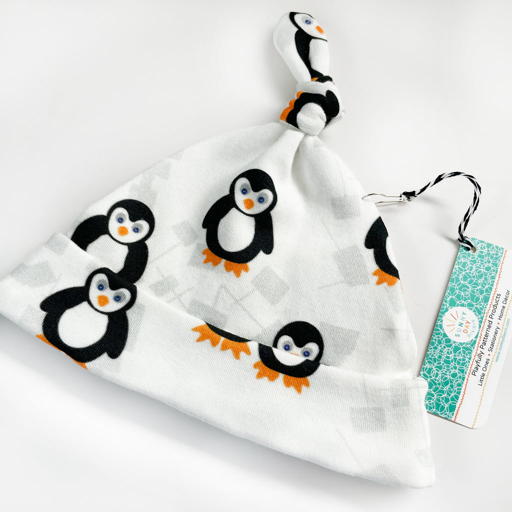 A tilted view of an adorable black, white, blue and orange organic cotton baby hat made from a penguin printed fabric by Sunny Day Designs and packaged with a branded Sunny Day Designs logo hang tag
