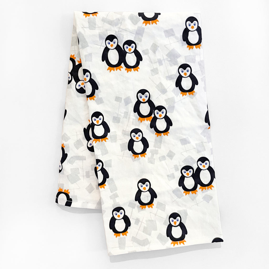 A playful penguin print linen/cotton tea towel in black, yellow, gray & white is shown unpackaged and folded in half on a white background. This cute arctic animal ea towel is made in the USA by Sunny Day Designs and is the perfect way to decorate your kitchen for winter, Christmas or the holiday season!