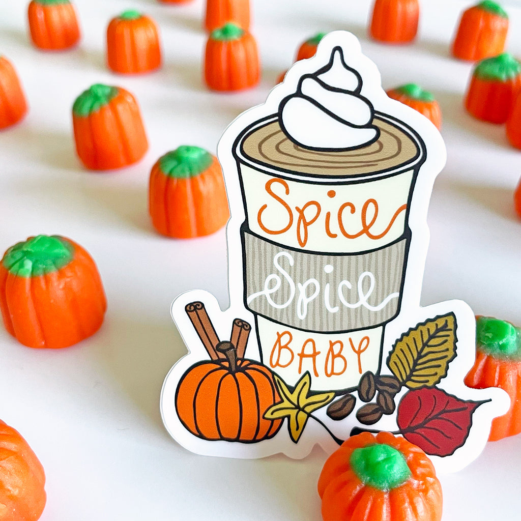 Pumpkin Spice Spice Baby vinyl sticker by Sunny Day Designs, featuring a pumpkin spice latte with whipped cream and fall accents like a pumpkin, cinnamon sticks, autumn leaves, coffee beans, and a star anise, displayed with candy pumpkins in the background.