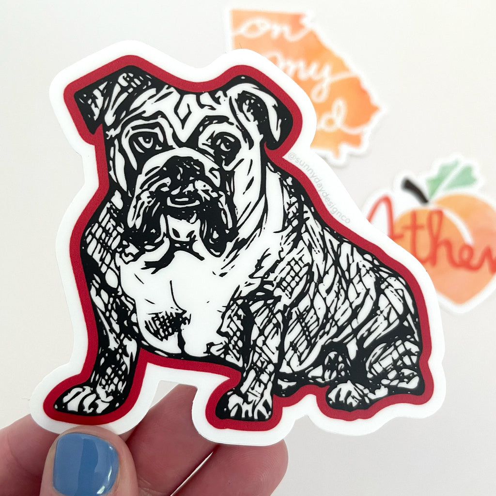 A black and Red Bulldog themed vinyl sticker is shown held in a hand with 2 other UGA and/or Georgia themed stickers in the background