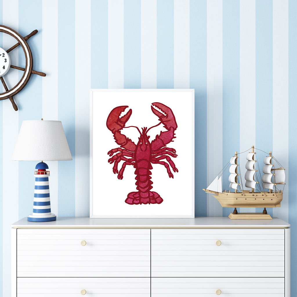 A watercolor illustration of a red lobster on a white background is shown framed and resting on a white dresser while leaning up against a blue/white vertical striped wall in a nautical-themed children's bedroom. This whimsical 8x10 coastal lobster wall art print coordinates with the other seaside decor shown in the room, such as a diorama of a ship, a red/white/blue lighthouse-shaped table lamp, and a wooden ship wheel-shaped wall clock. Art print is designed by Sunny Day Designs & is made in the USA.