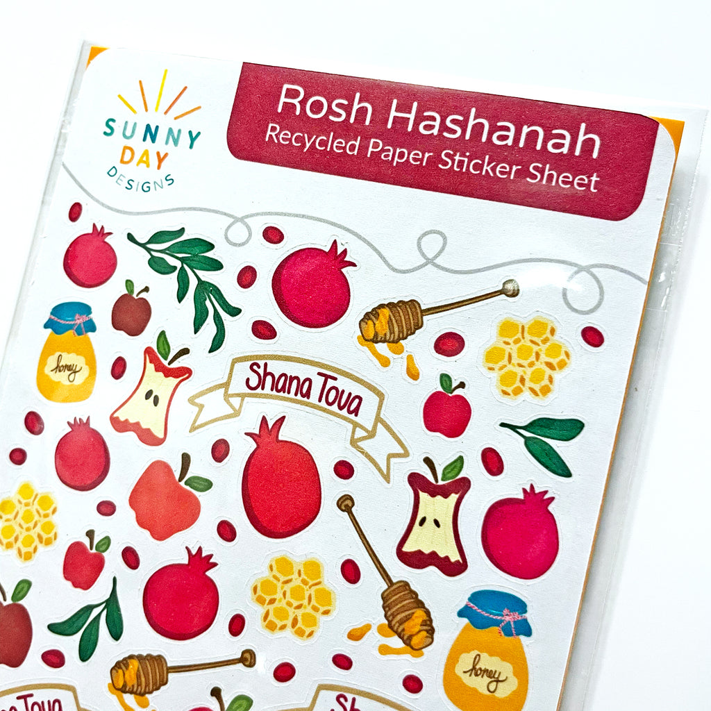 A close up view of our packaged Rosh Hashanah Jewish new year holiday sticker sheet, which features 71 small uncoated recycled paper stickers in the following designs: pomegranates, leaves, apples, honey, honeycomb, and Shana Tova banners. 