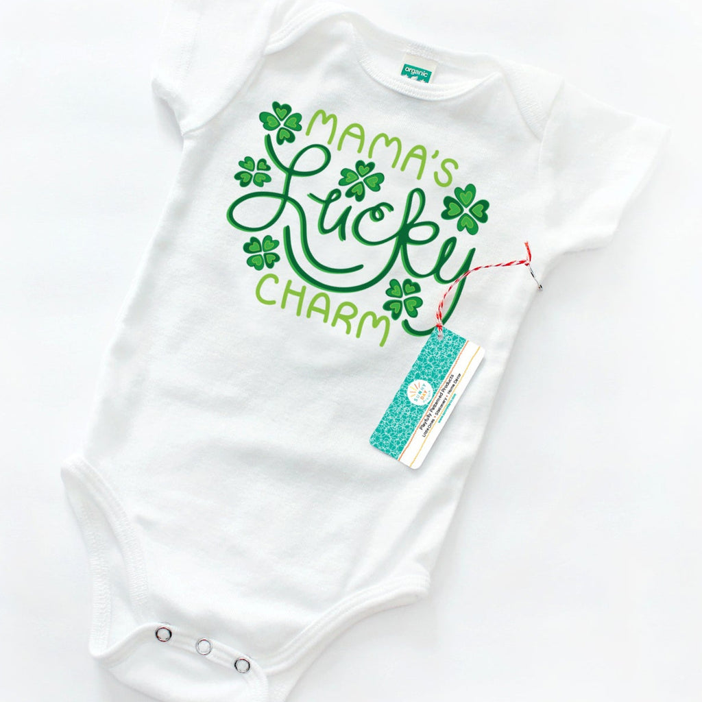 a green and white four-leaf clover Irish-themed organic cotton baby onesie is shown tilted on a white background and is printed with the words "Mama's Lucky Charm" and 6 green shamrock motifs