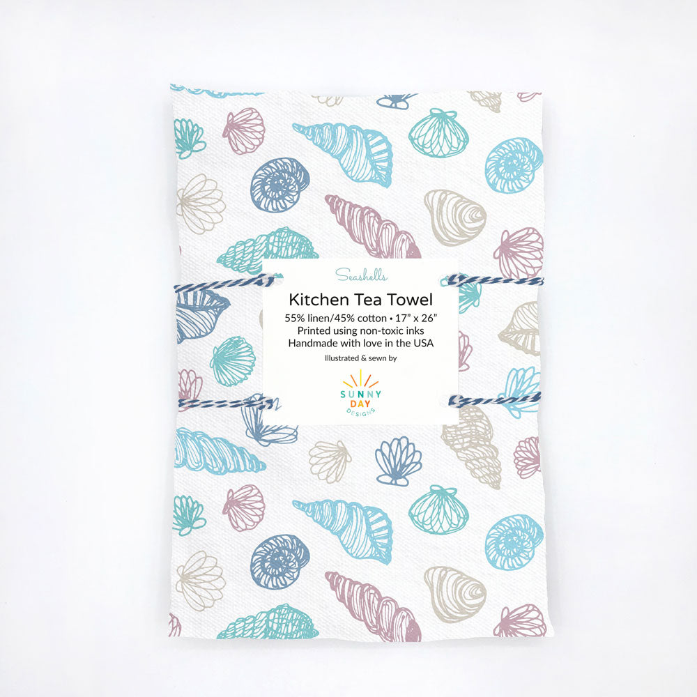 Packaged 'Seashells' kitchen tea towel by Sunny Day Designs, featuring a delicate seashell pattern in soft pastel tones of blue, teal, and taupe on a white background. The towel is made from a 55% linen and 45% cotton blend, printed with non-toxic inks, and is handcrafted in the USA.