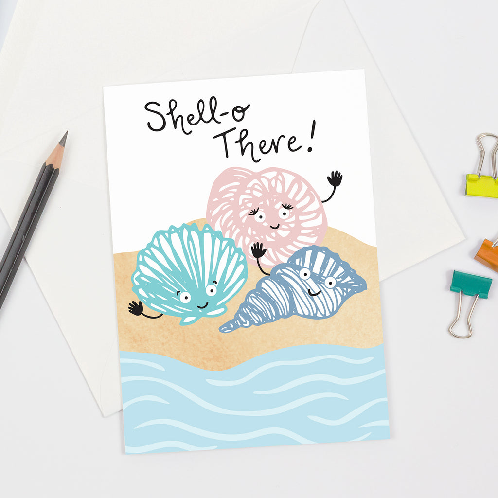 Greeting card with three smiling seashells in green, pink, and purple pastel colors on a sandy shore with gentle waves. The text 'Shell-o There!' is displayed above the seashells. The card is shown with a white envelope, pencil, and yellow, orange and green binder clips arranged around it.