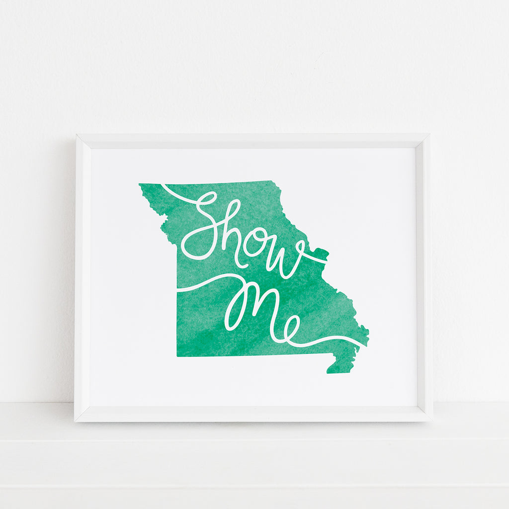 Framed 'Show Me' Missouri art print in green by Sunny Day Designs. The artwork features the state of Missouri in a fresh green watercolor style with 'Show Me' elegantly scripted across. Displayed in a white frame against a minimalist background, adding a vibrant touch to any decor.