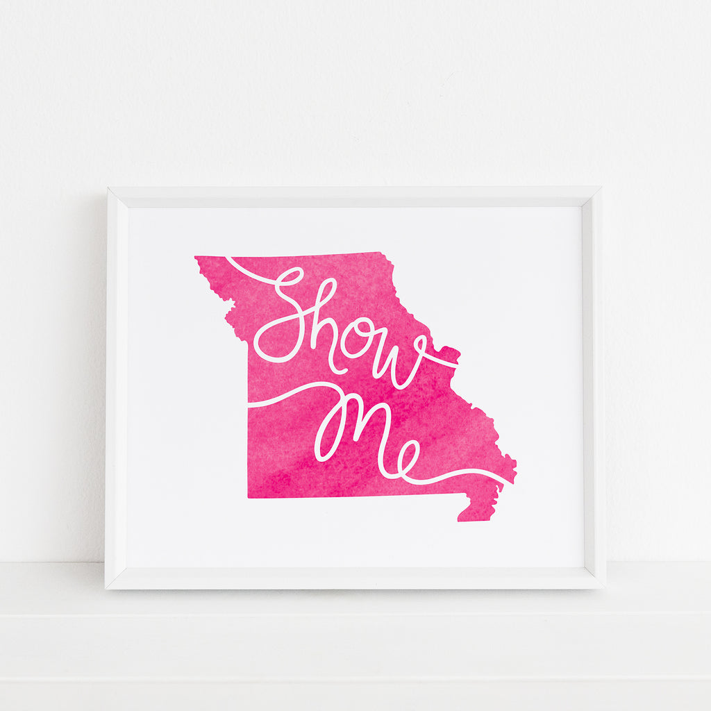 Framed 'Show Me' Missouri art print in magenta by Sunny Day Designs. The artwork features the state of Missouri in a bold pink watercolor style with 'Show Me' scripted across. Displayed in a minimalist white frame, it rests against a white background, adding a bright pop of color to the setting.