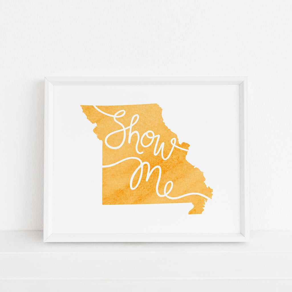 Framed 'Show Me' Missouri art print in yellow by Sunny Day Designs. The artwork features the state of Missouri in a vibrant yellow watercolor style with 'Show Me' scripted across. Displayed in a clean white frame against a white background, it adds a warm and cheerful accent to the setting.