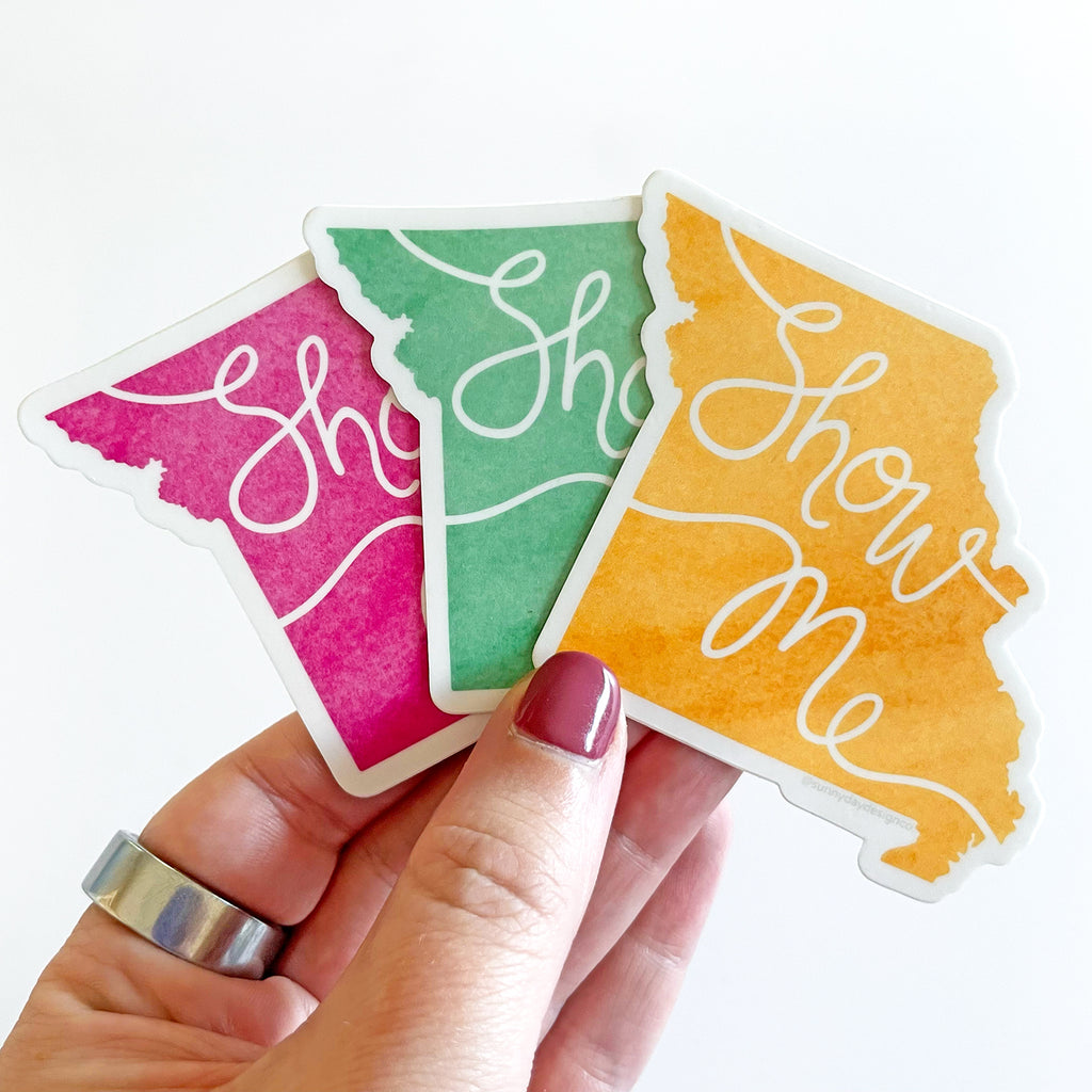 A hand is shown holding 3 colorful watercolor Missouri state-shaped vinyl stickers by Sunny Day Designs; 1 sticker is magenta pink, 1 sticker is green, and 1 sticker is golden yellow.