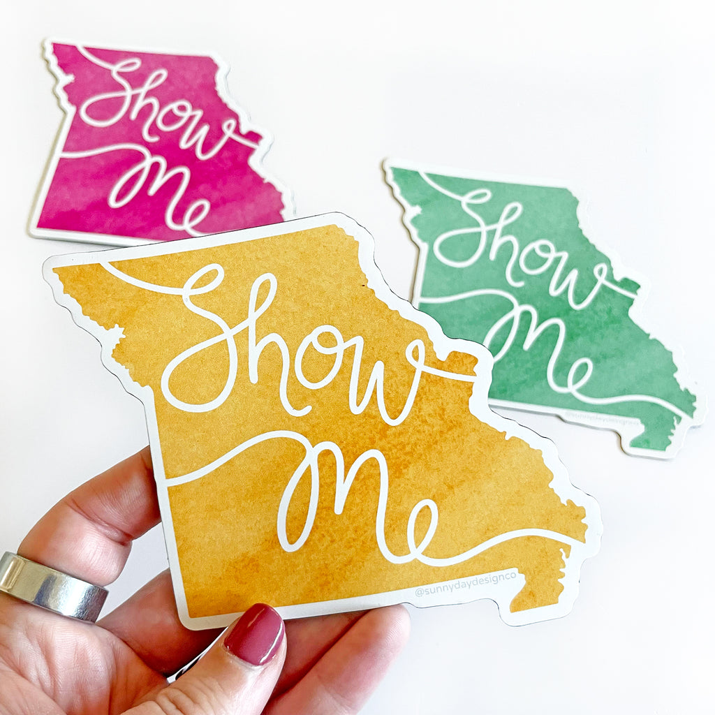 A hand is shown holidng a golden yellow colored Missouri state-shaped vinyl magnet in front of a white background with hand lettered white script on the magnet that says "Show Me" as a reference to Missouri's state nickname: "The Show Me State". In the background you can also see the same magnet in 2 other colorway options: magenta pink and green. All 3 magnets are joyfully designed and made in the USA by Sunny Day Designs.