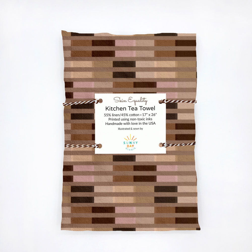 Packaged Skin Equality kitchen tea towel featuring a pattern of skin tone-inspired blocks in shades of beige, brown, and tan. The tea towel is made from a linen and cotton blend, printed using non-toxic inks, and handmade with love in the USA. The towel is wrapped with string, emphasizing its thoughtful and inclusive design, perfect for adding a meaningful touch to any kitchen.