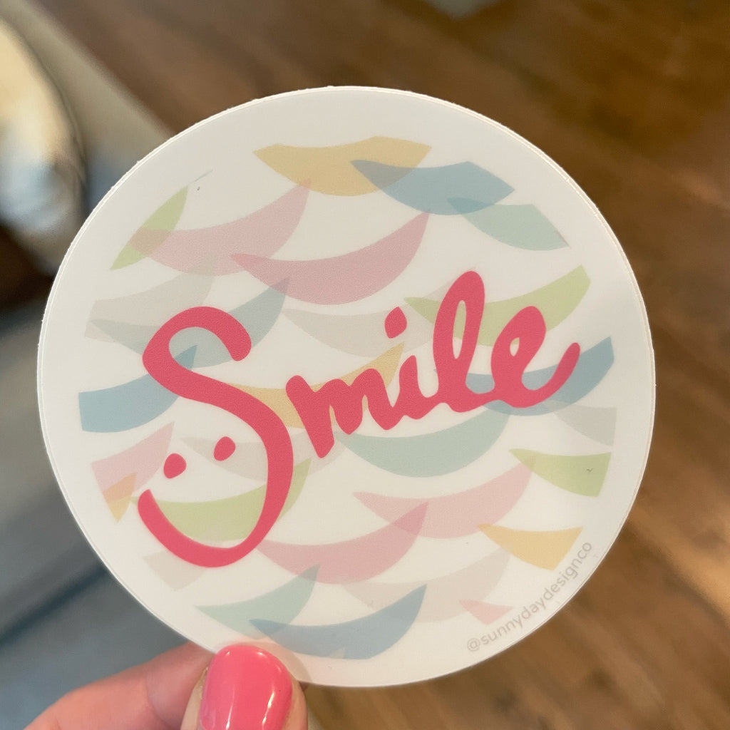A colorful, round waterproof laptop sticker is shown held in someone's fingers with the word "Smile" hand lettered in pink in a clever way that includes 2 eye "dots" above the lower bend of the "S" letter so that it looks like there is a smiley face in the S-shape. Behind the handlettered "Smile" text there are pastel-colored smile crescent shapes layered on top of one another to create a colorful & fun patterned background. This adorable positivity sticker is joyfully made in the USA by Sunny Day Designs.