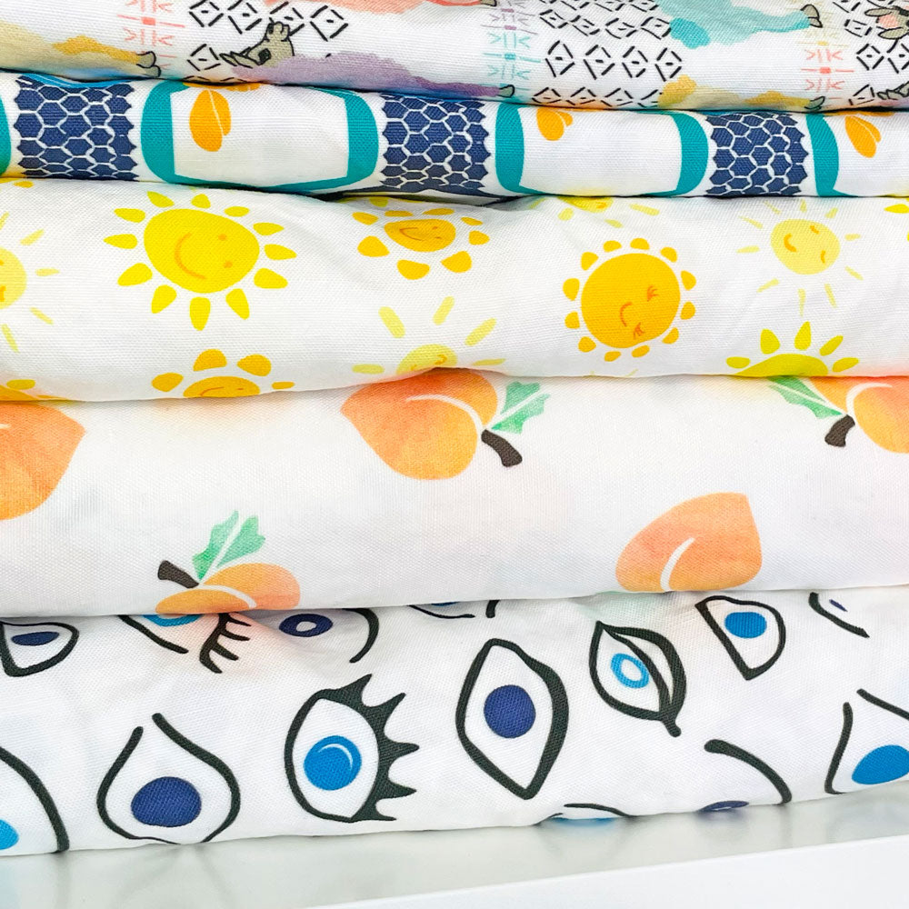 3 colorful printed fabrics (featuring smiling suns, watercolor peaches, and evil eye illustrations by Sunny Day Designs) are folded and stacked on top of each other