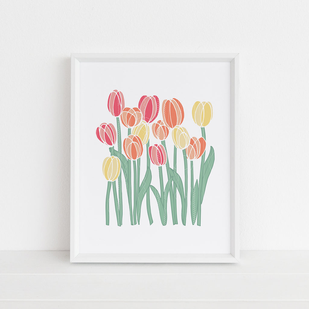 A colorful floral art print is shown inside a white picture frame - the print design features yellow, orange, and pink tulip blooms on a white background with a white stitched tetture overlay. Artwork is by Sunny Day Designs.