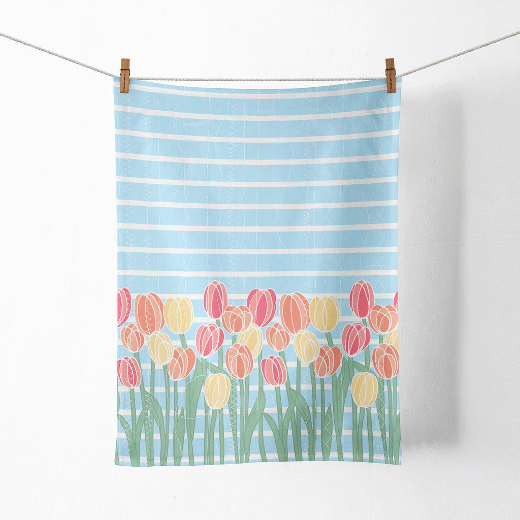 A fully open view of a colorful, floral printed kitchen tea towel by Sunny Day Designs, which is shown hanging from clothespins against a white wall.