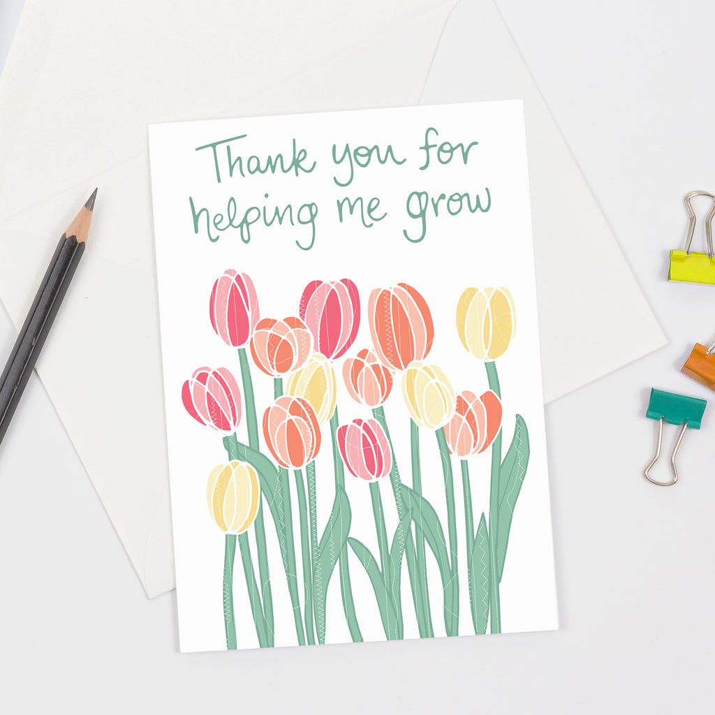 A floral themed gratitude greeting card that is perfect to give to mothers, teachers, and more - featuring colorful orange, pink, and yellow tulips against a white background with green handlettered text (above flowers) that says "Thank you for helping me grow".