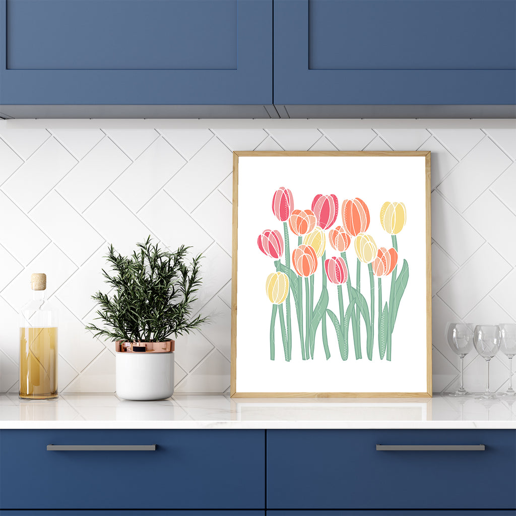 A beautiful, framed floral wall art print (the Stitched Tulips 8x10 watercolor art print by Sunny Day Designs) is shown leaning against white subway tile on top of a white kitchen counter with blue cabinets. This colorful flower-themed wall art features a stitched/sewn texture on top of a colorful illustration of 13 pink, orange and yellow tulips that evokes the freshness and beauty of springtime.