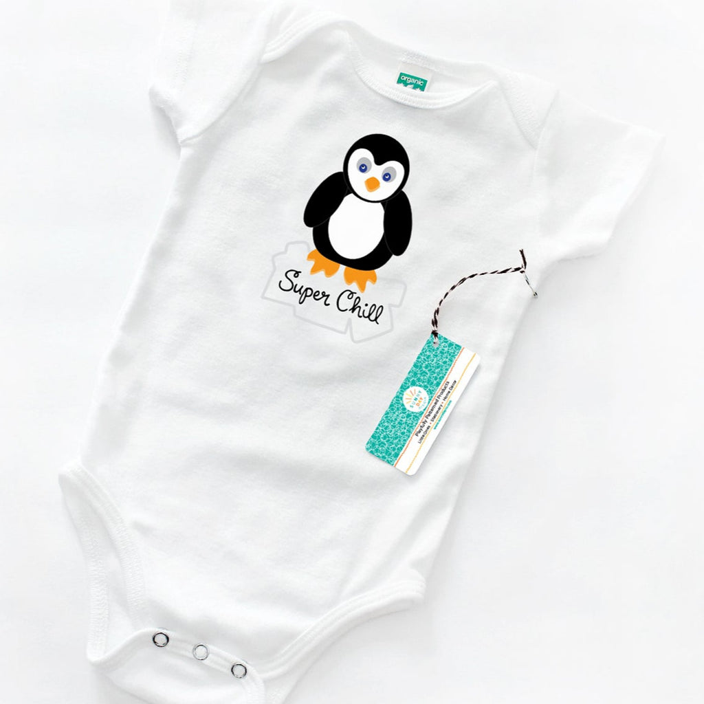 An organic cotton white baby onesie featuring a cute cartoon penguin standing on an iceberg with the phrase “Super Chill” in playful hand lettered text. This made in the USA baby onesie is laid flat on a white background with an attached Sunny Day Designs hang tag.
