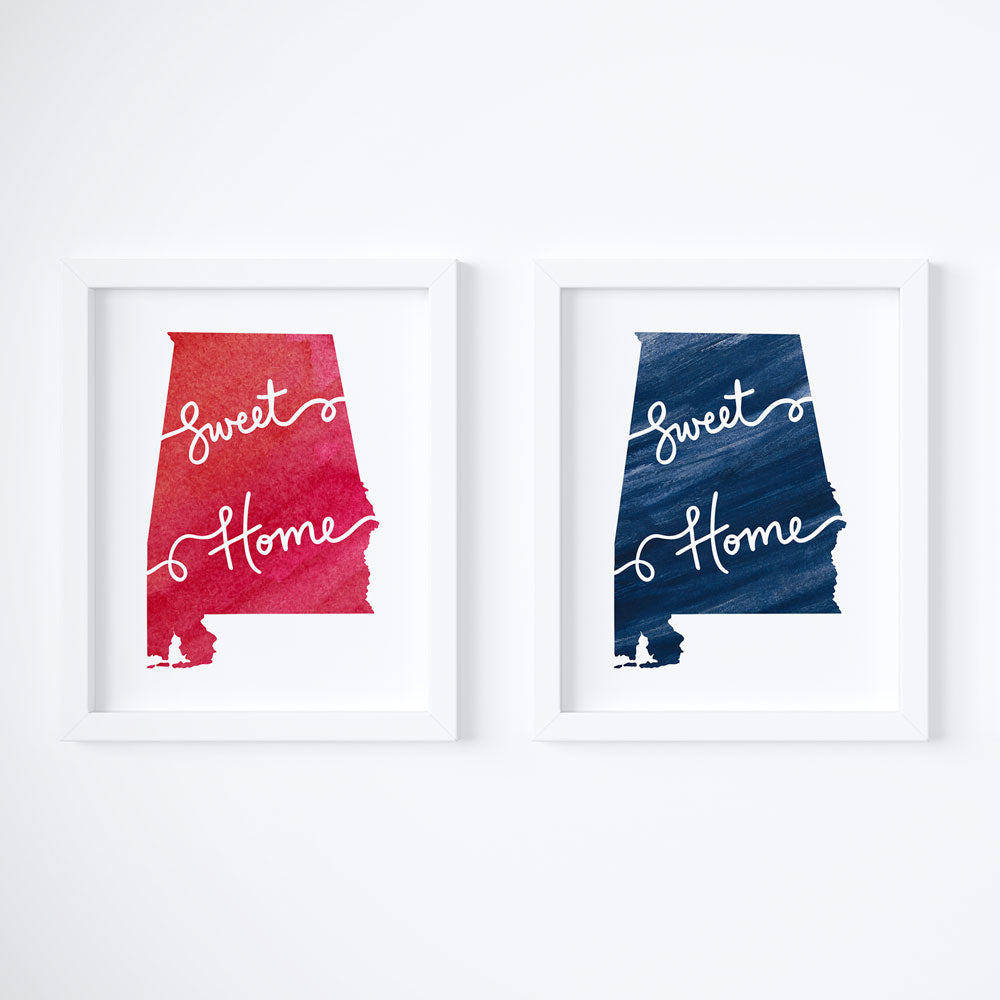 2 Alabama state-themed 8x10 art prints are shown framed inside a white picture frame and hung side-by-side on a white wall. The left AL wall art features a Crimson Tide red watercolor Alabama state shape with hand lettered white script that says "Sweet Home". The right Alabama art print features an Auburn Blue watercolor Alabama state shape with hand lettered white script that says "Sweet Home", with an ode to the song Sweet Home Alabama.