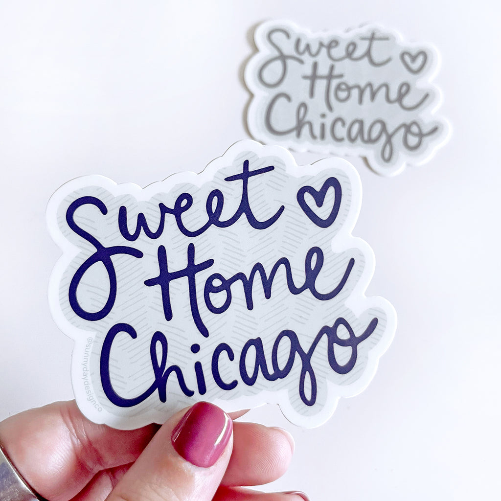 A hand is shown holding a cute Chicago-themed vinyl sticker that says "Sweet Home Chicago" in a navy blue and gray colorway, while a 2nd Charcoal Gray color option of the same "Sweet Home Chicago" sticker design is shown in the background, laying on a white background. These cute city-themed stickers make great gifts for Chicagoans & are also great souvenirs to remember a trip to Chicago. Both stickers are made in the USA and designed in Madison, WI, by Sunny Day Designs.