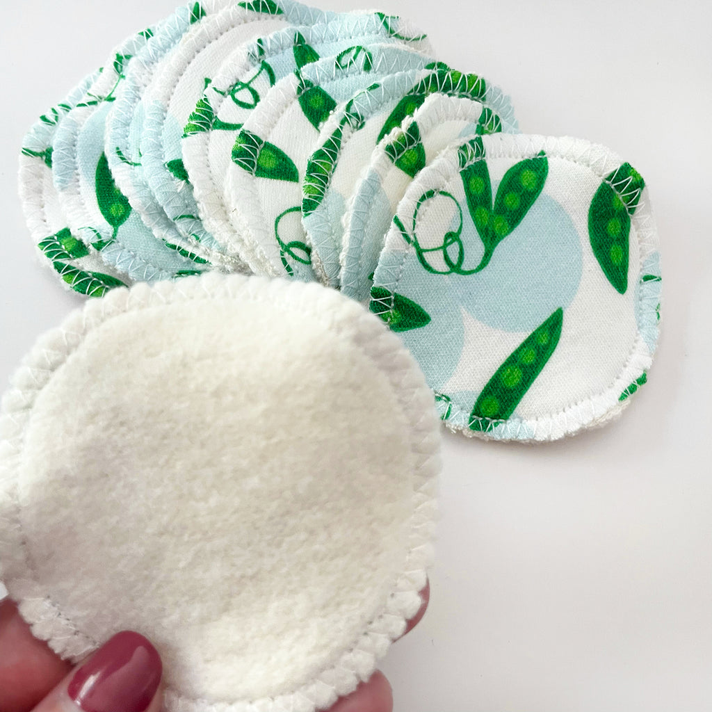 A set of 10 Sweet Pea printed organic cotton facial rounds are shown fanned out with the printed green/blue vegetable print visible and 1 hand holding a wipe and showing the tan organic cotton fleece fabric on the back side of each wipe.