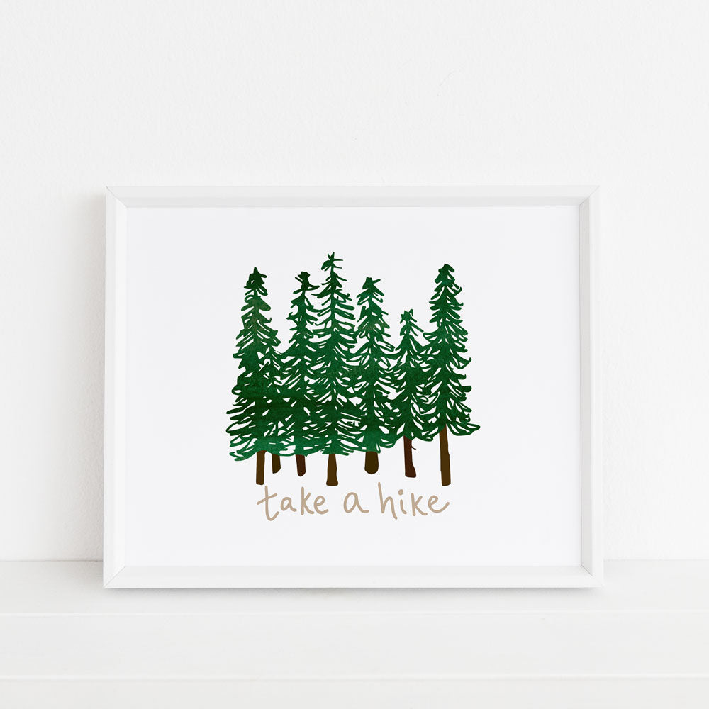 A forest and tree themed 8x10 art print framed inside a white picture frame features 7 green watercolor trees and hand lettered "take a hike" text. This nature-themed artwork was created by Sunny Day Designs in Madison, WI, and is joyfully made in the USA.