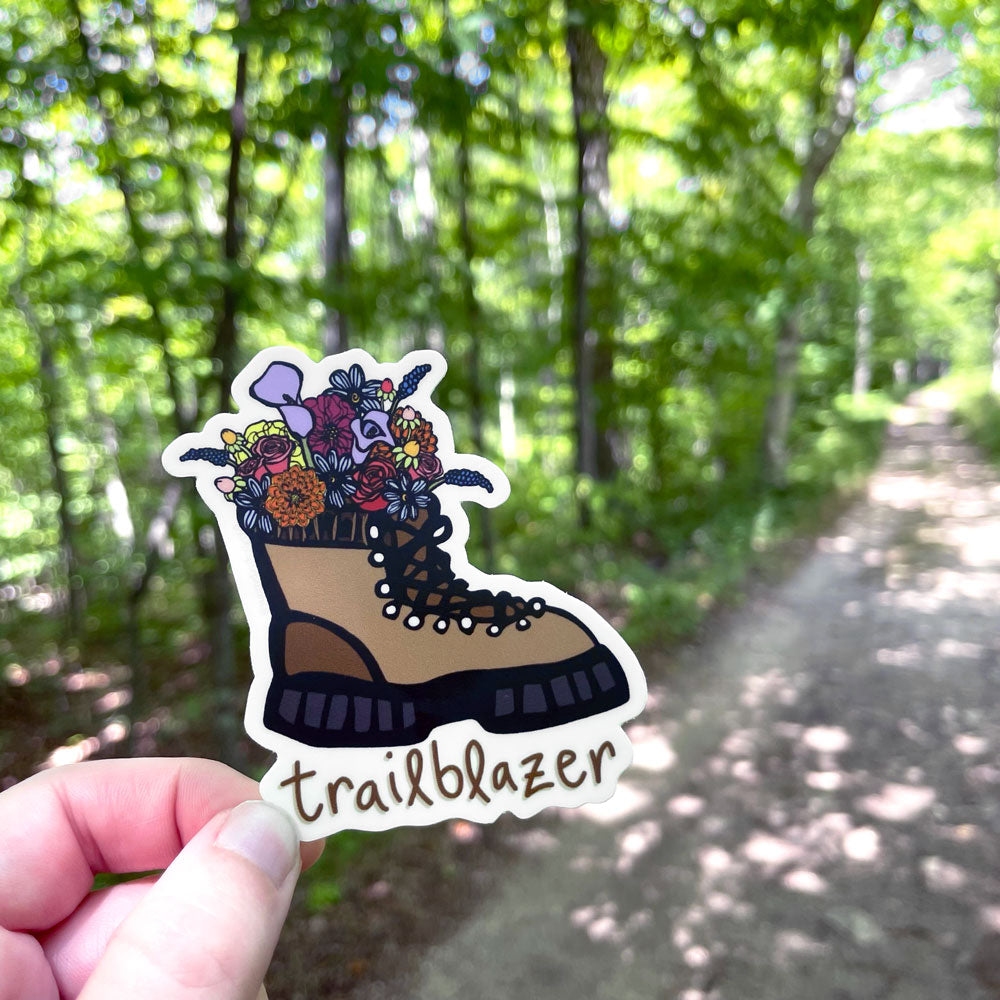 A hiking boot-themed vinyl sticker by Sunny Day Designs with flowers sprouting inside the top of the boot and hand lettered "Trailblazer" text shown below it is shown being held in 1 hand in front of a hiking trail path.