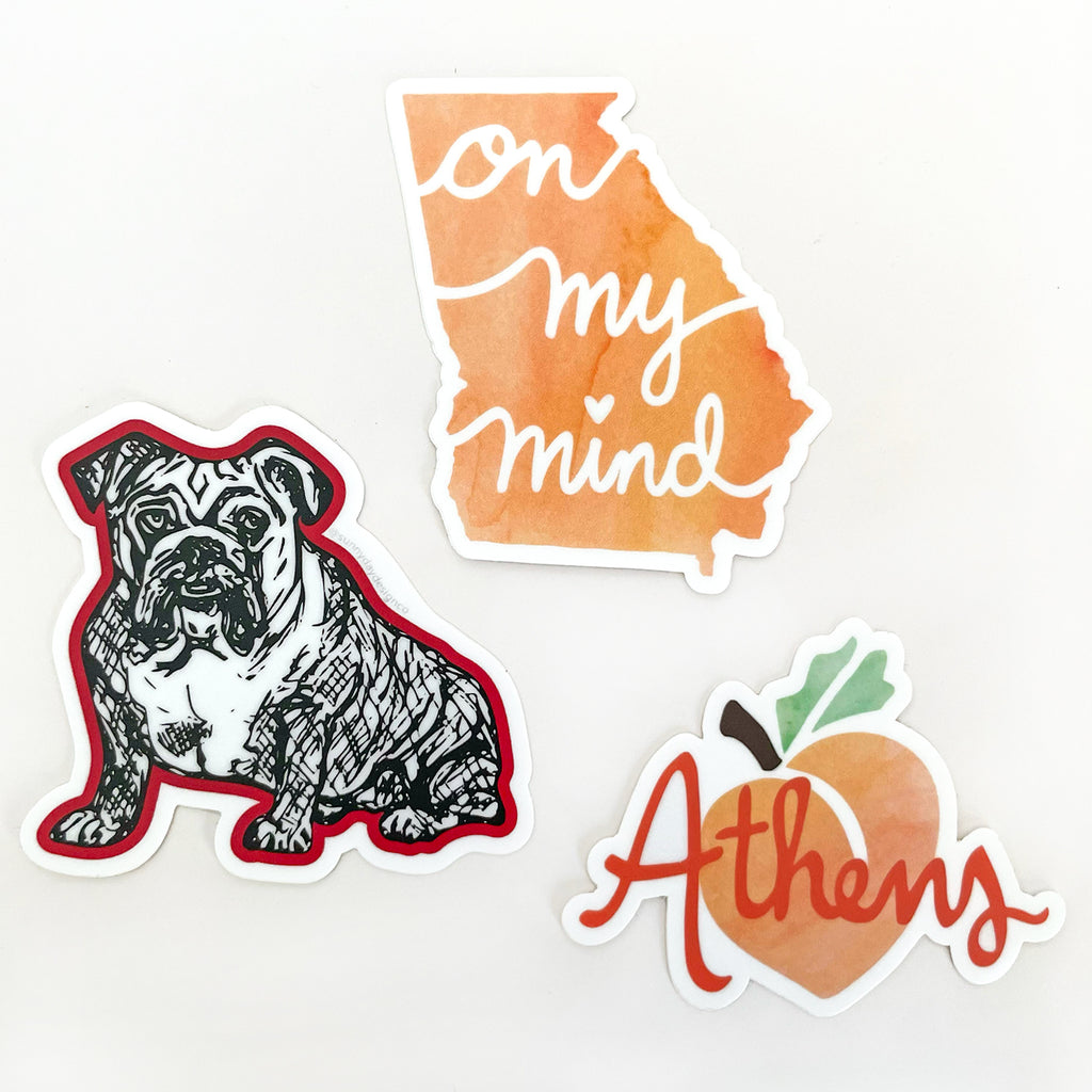 A curated gift set of 3 UGA stickers is shown on a white background and includes 1 Red Bulldog vinyl sticker, 1 orange peach watercolor Georgia On My Mind state-shaped vinyl sticker, and 1 Athens GA Peach vinyl sticker. All 3 die cut stickers are designed by Sunny Day Designs and made in the USA - a unique small gift for UGA fans, alumni, students and graduates and also makes a fun Athens, GA souvenir!