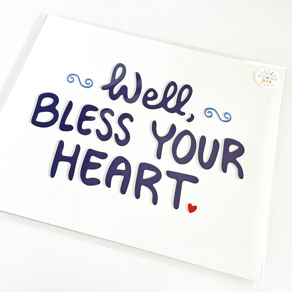 Close-up view of the 'Well, Bless Your Heart' 8x10 art print by Sunny Day Designs, featuring bold navy blue lettering with a small red heart and decorative blue swirls, packaged in a clear protective sleeve with Sunny Day Designs branding.