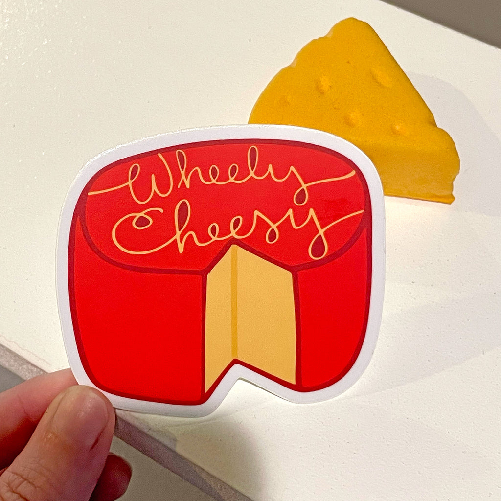 A cute red, yellow and white vinyl sticker by Sunny Day Designs in the shape of a wheel of cheese with a slice cut out of it and illustrated script on the top that says "Wheely Cheesy". This Wheely Cheesy sticker is held in a hand in front of a white table with a small yellow cheese wedge shown in the background.